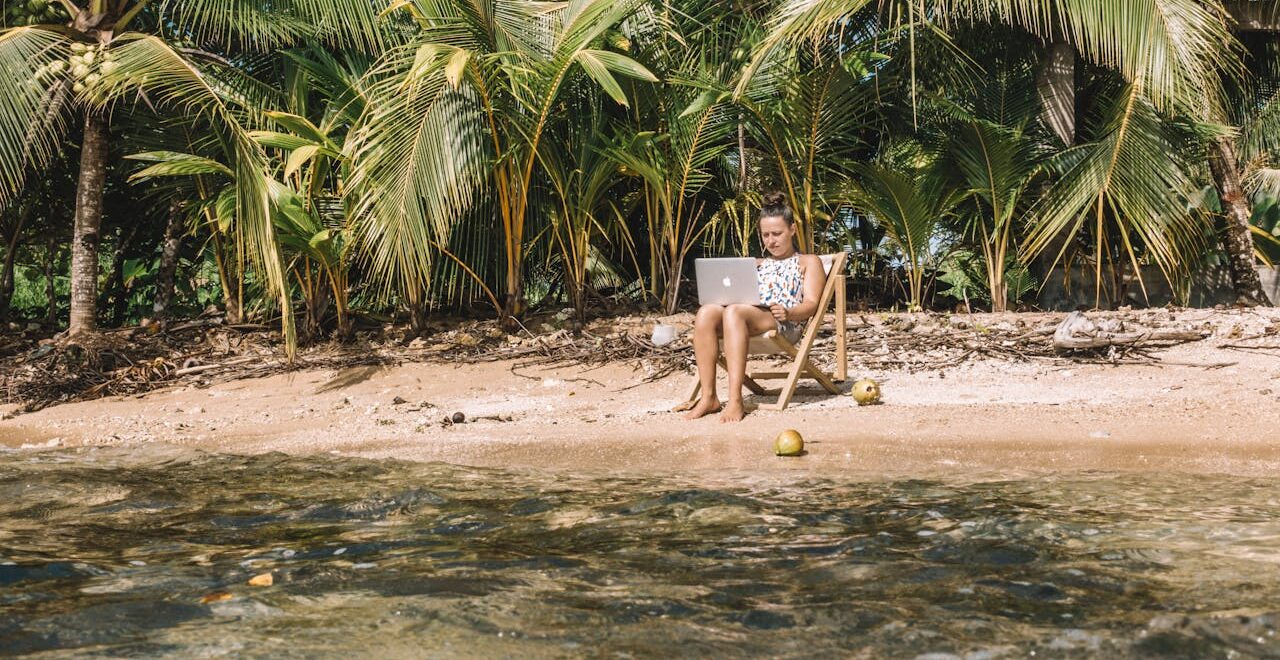 Woman working remotely with a laptop on a tropical beach in Panama surrounded by lush green palm trees.